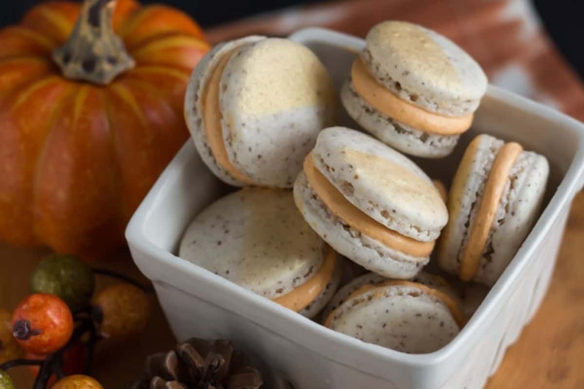 A white square dish is filled with several macarons that have light-colored shells and orange filling. The dish is placed on a wooden surface alongside Thanksgiving-themed decorations, including a small pumpkin, pine cones, and assorted berries.