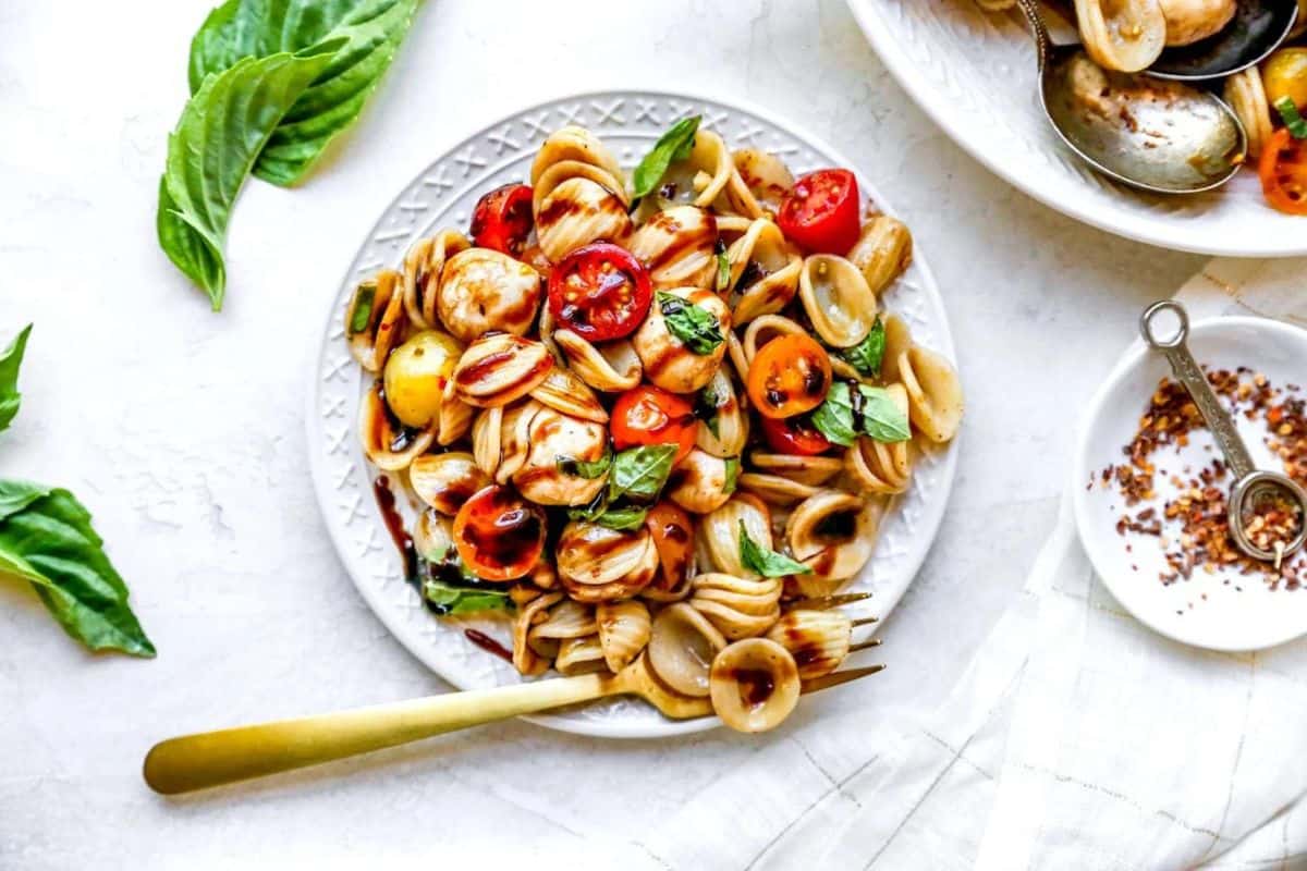A white plate of orecchiette pasta topped with cherry tomatoes, mozzarella balls, and fresh basil leaves sits on a table. A fork rests on the plate's edge. Fresh basil leaves are scattered around, and a small dish with a spoon and red pepper flakes is next to the plate.