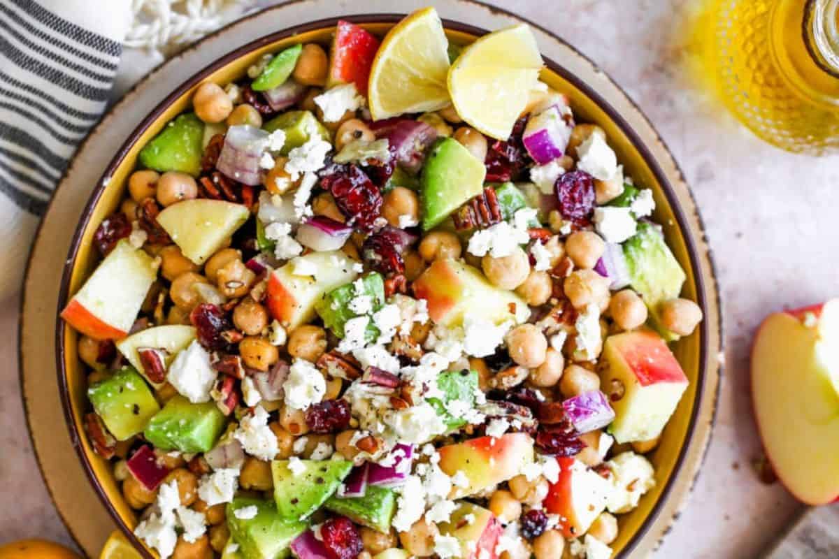 A vibrant salad with chickpeas, avocado, diced apples, red onion, cranberries, feta cheese, and pecans, garnished with lemon wedges. The salad is in a wooden bowl on a light-colored surface, accompanied by a striped cloth and a part of a sliced apple beside it.