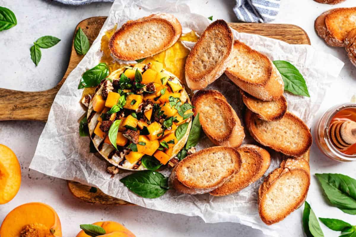 A wooden board holds a serving of crostini, with toasted bread slices surrounding a topping of diced peaches, basil leaves, and honey drizzle on a white parchment paper. Fresh peach slices, basil leaves, and a honey dipper are arranged around the board.