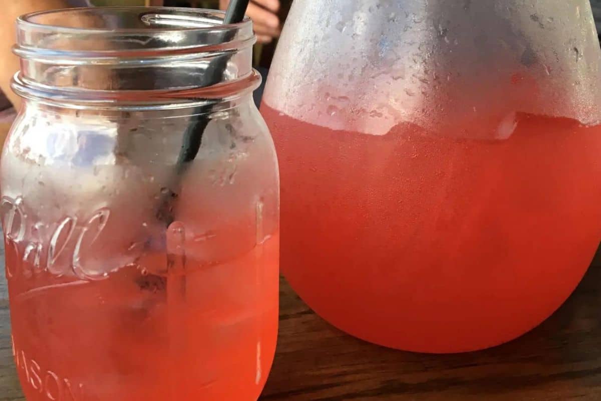 A glass jar with a black straw, filled with iced pink lemonade, sits beside a large, frosty pitcher containing more pink lemonade. Both containers are on a wooden table.