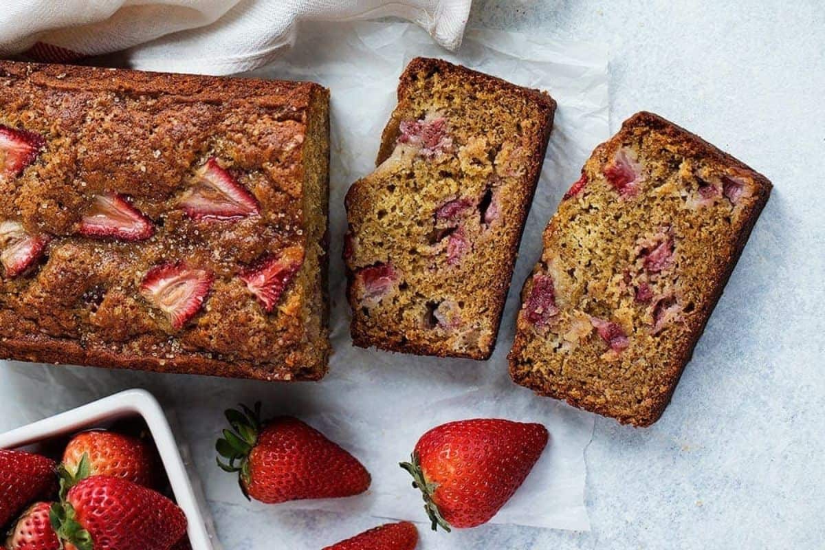 A loaf of strawberry banana bread is partially sliced on a white parchment paper, revealing two thick slices filled with chunks of strawberries. Fresh strawberries are scattered around the bread, and a white cloth is partially visible in the background.