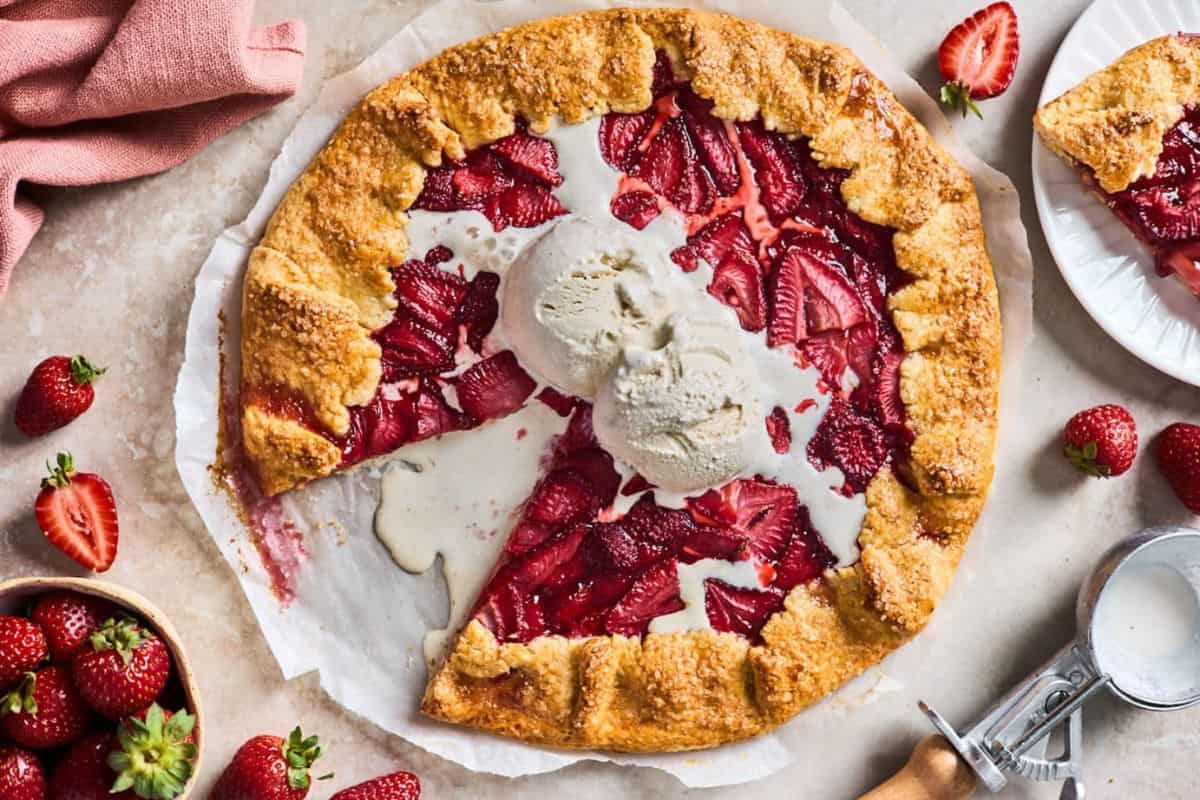 A rustic strawberry galette with a golden, flaky crust sits on a parchment-lined surface. A scoop of vanilla ice cream melts on top of the filling. Fresh strawberries and a slice of the galette are nearby, along with a pink napkin and an ice cream scoop.