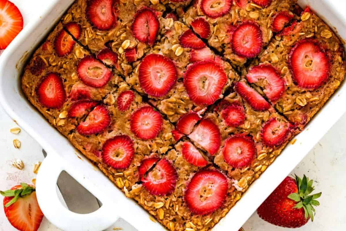 A baked oatmeal dish topped with fresh strawberry slices is cut into square portions inside a white ceramic baking dish. The oatmeal is golden brown and garnished with whole oats. Fresh strawberries are placed around the dish for decoration.
