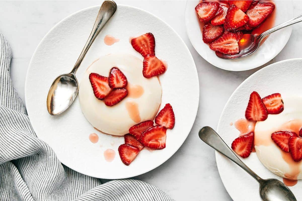 Three white plates, each with a serving of panna cotta topped with sliced strawberries and drizzled with strawberry syrup. Two plates have silver spoons resting beside the dessert. A gray and white striped napkin is partially visible on the left.