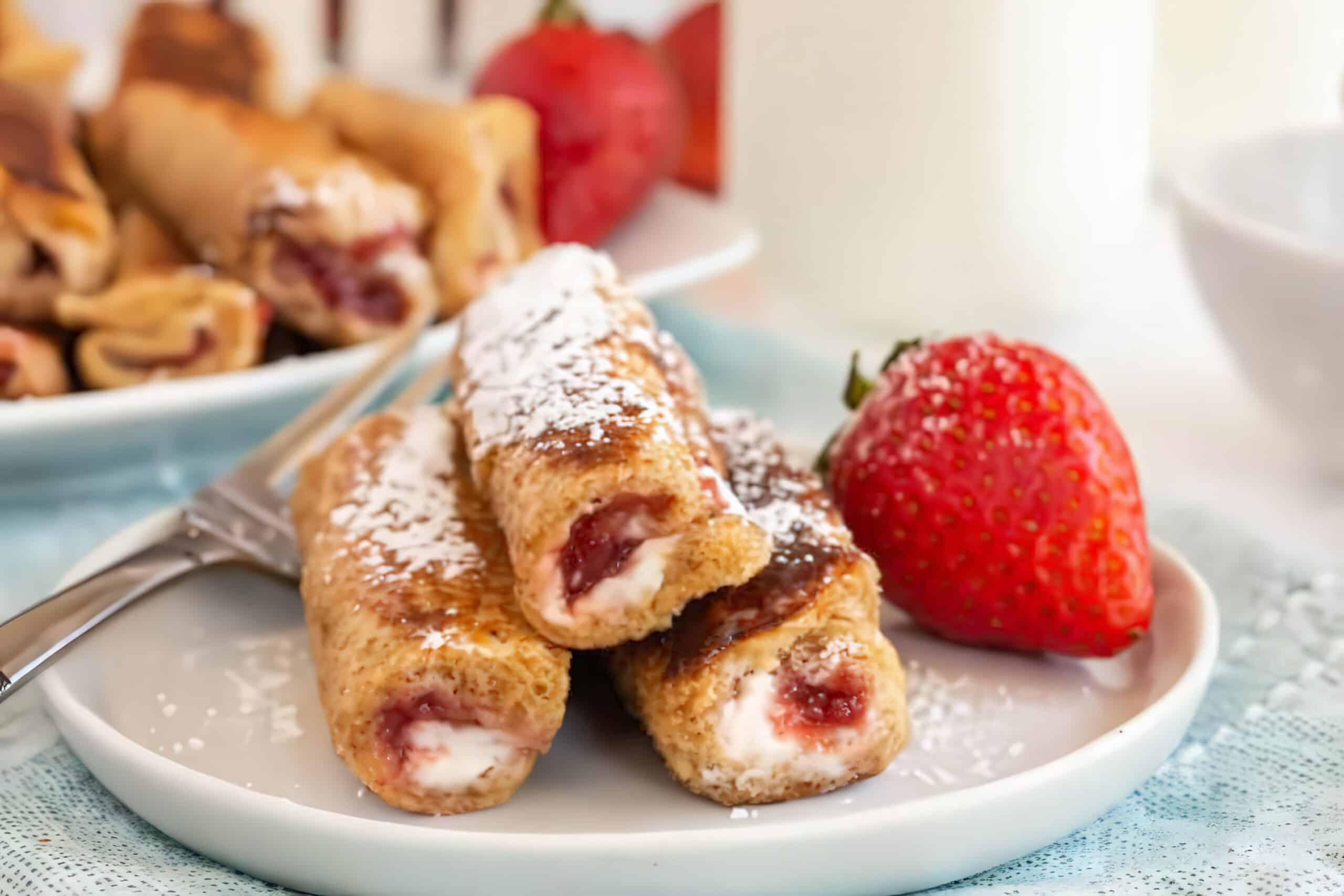 A plate with three strawberry and cream cheese French toast roll-ups dusted with powdered sugar, garnished with a fresh strawberry. In the background, another plate of roll-ups and a glass of milk are visible.