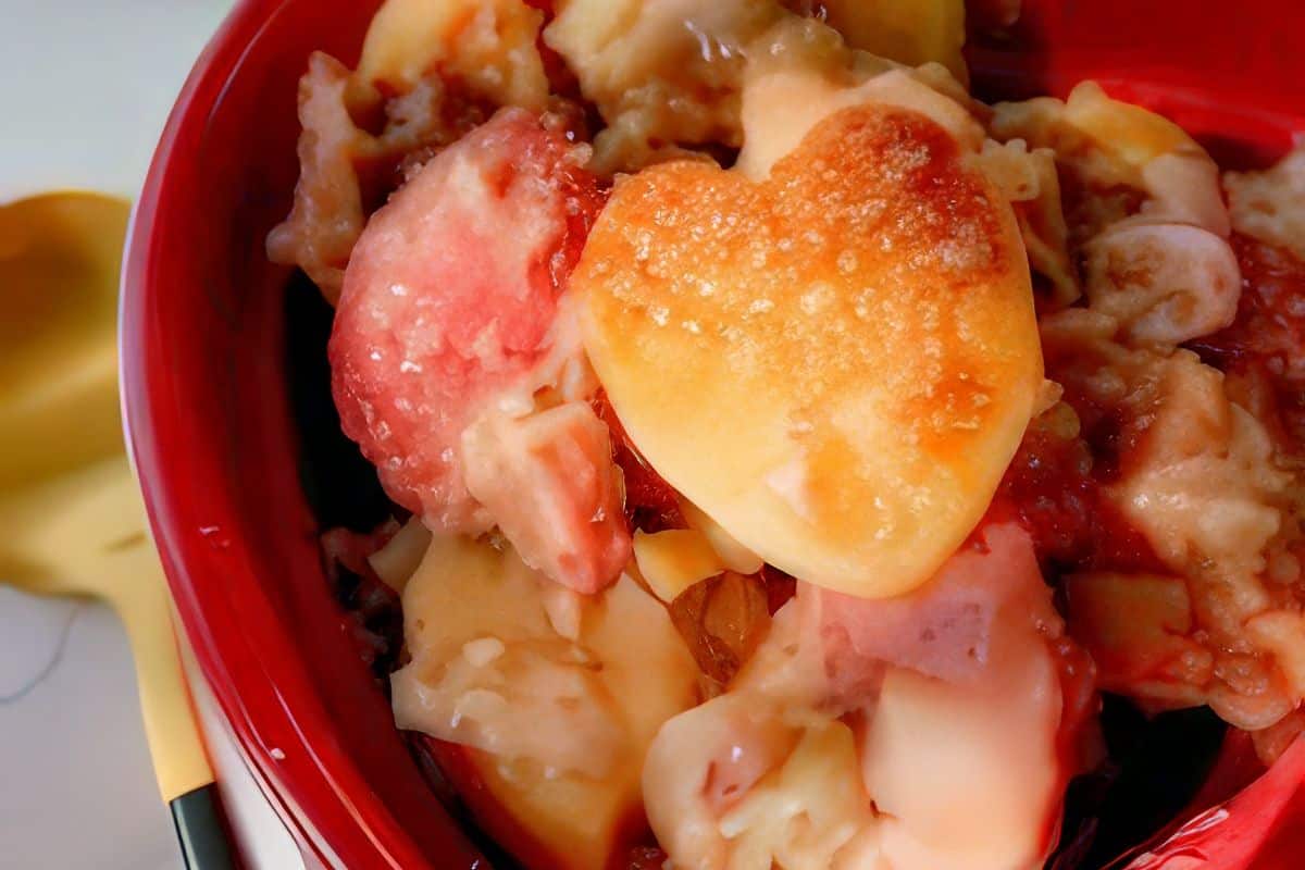A close-up of a dish featuring heart-shaped pieces of fruit, possibly apples or peaches, mixed with other ingredients in a red bowl. The mixture appears to be a baked dessert, such as a fruit cobbler or pie, with a spoon visible to the left.