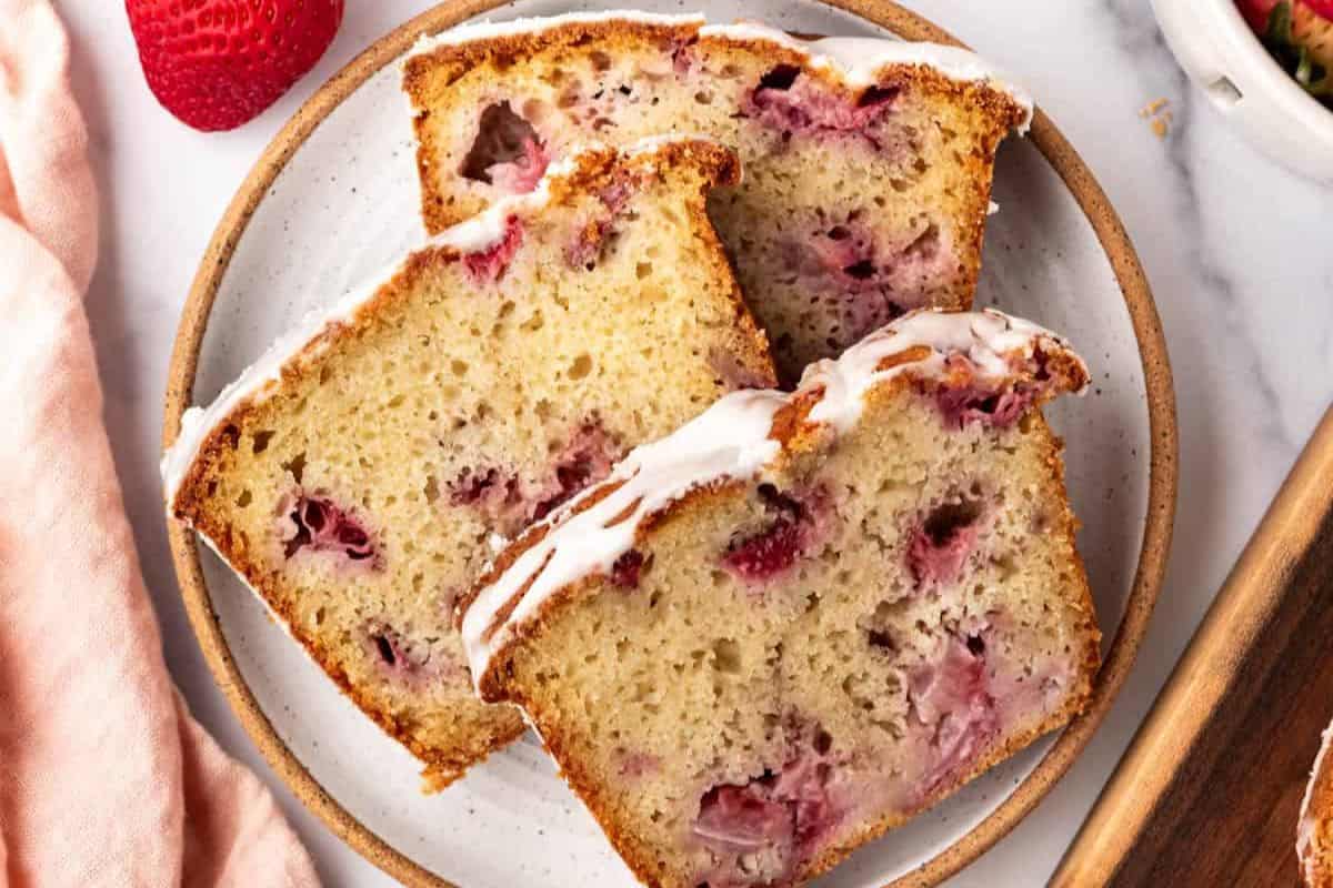 Two slices of strawberry bread with a white glaze on top, served on a round plate. The bread has visible chunks of strawberries and a lightly browned crust. A fresh strawberry is in the background, along with a pink cloth and a wooden cutting board.