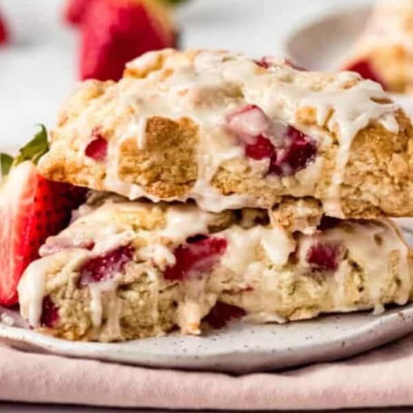 Two strawberry scones stacked on a small white plate are topped with a drizzle of icing. Fresh strawberries are placed beside the scones, and another plate with a scone is in the background, set on a light pink cloth.