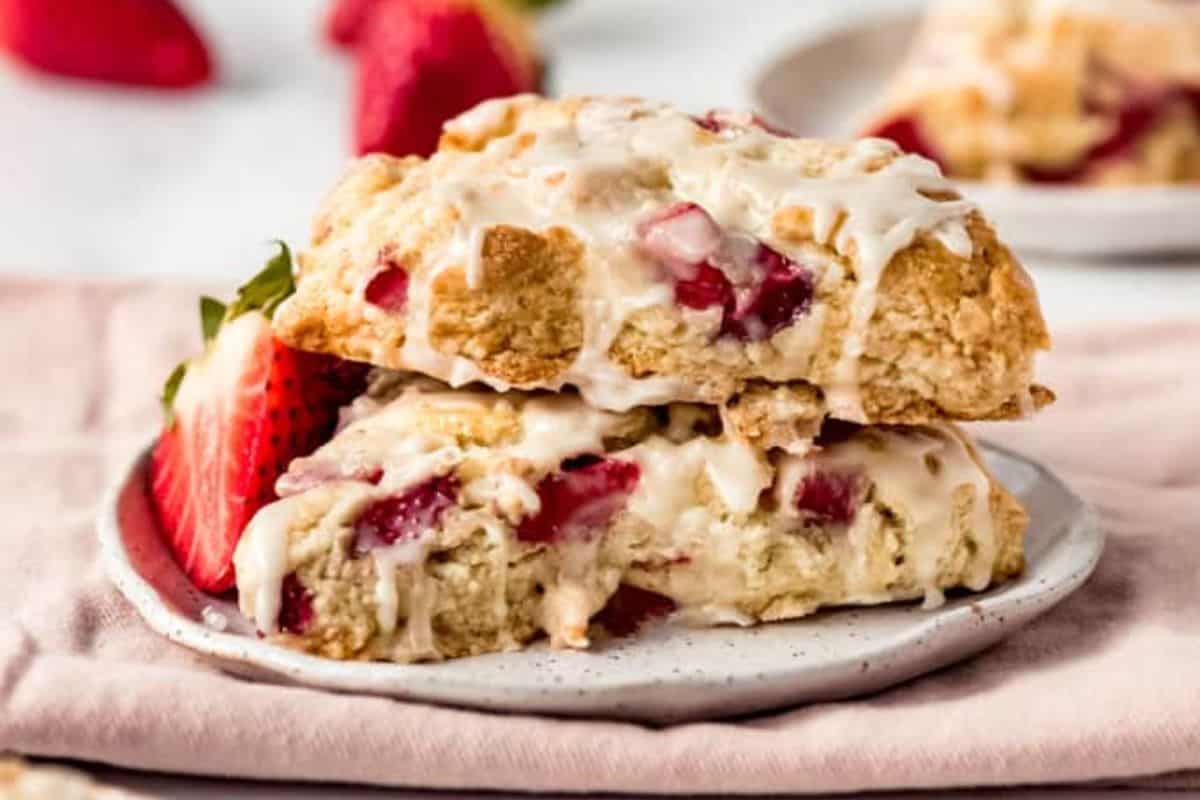 Two strawberry scones stacked on a small white plate are topped with a drizzle of icing. Fresh strawberries are placed beside the scones, and another plate with a scone is in the background, set on a light pink cloth.