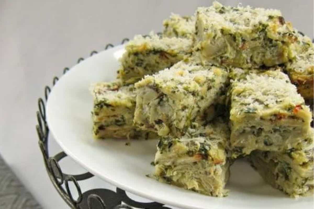 Square slices of savory bread pudding are arranged in a pyramid shape on a white plate with a decorative metal stand. The bread pudding is filled with herbs and possibly vegetables, topped with a light sprinkling of grated cheese or breadcrumbs.