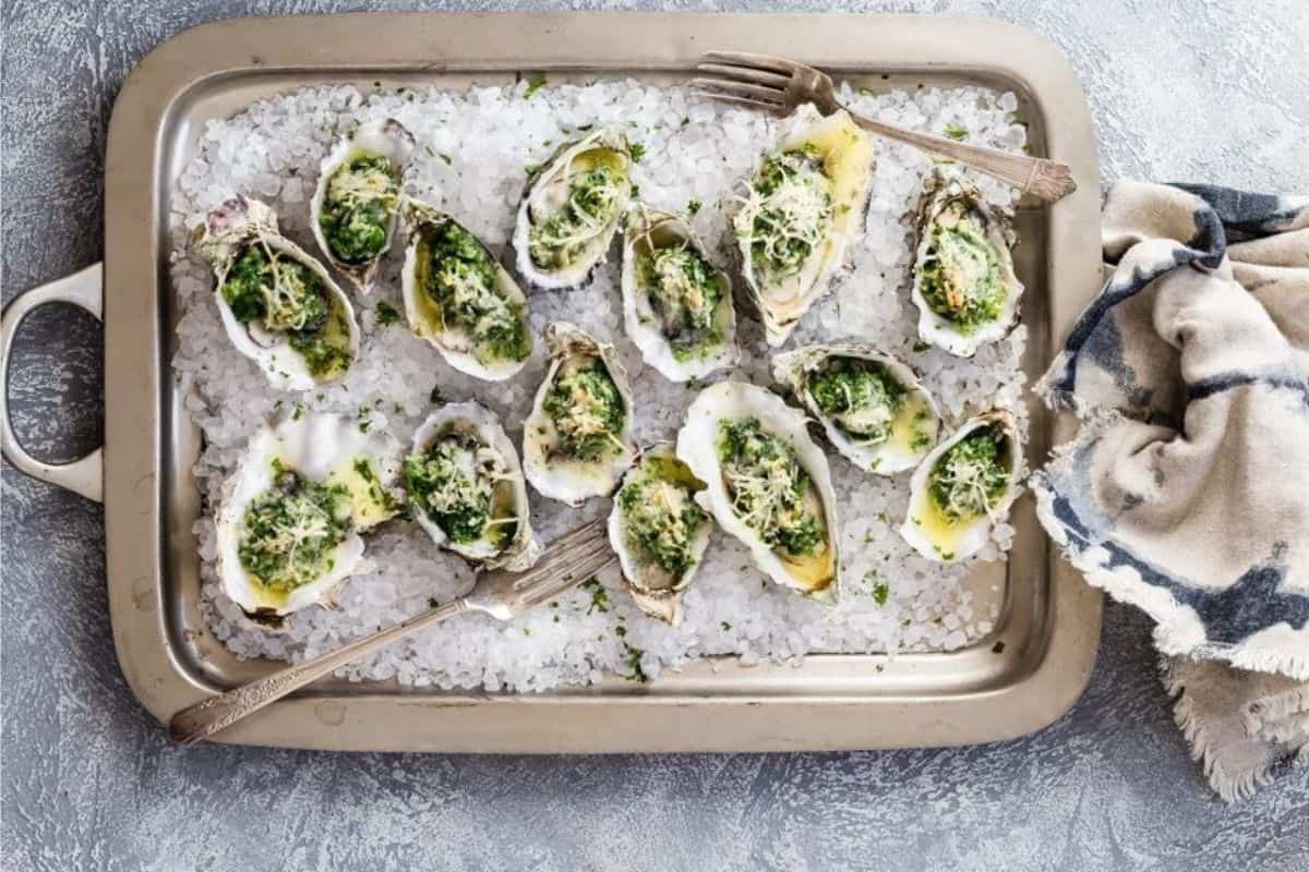 A rectangular tray filled with baked oysters on a bed of coarse salt. The oysters are topped with green herbs and melted cheese, and a fork is placed beside them. A cloth napkin is draped on the right side of the tray, creating a casual presentation.