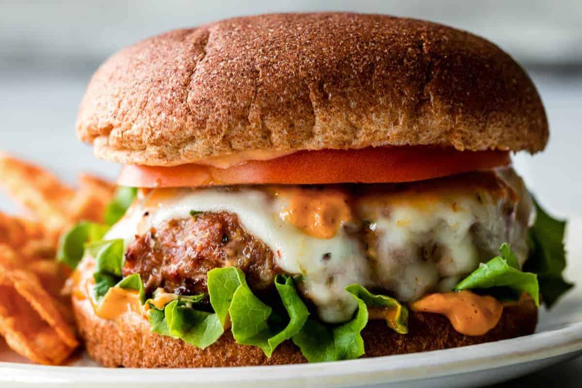 Close-up of a delicious burger with a whole wheat bun, melted cheese, leafy green lettuce, tomato slices, and a creamy sauce. Accompanied by a side of crispy fries. The burger is placed on a white plate.
