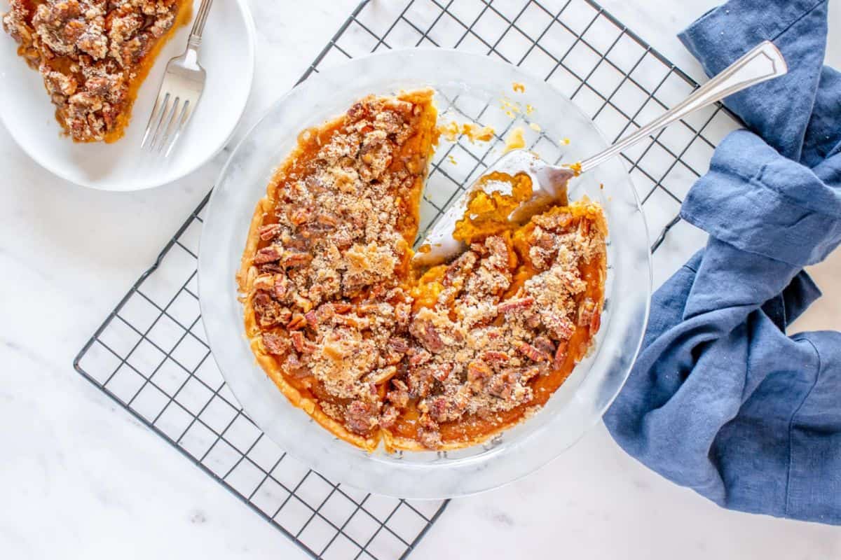 Top view of a partially eaten pecan-topped pie in a glass dish on a cooling rack. A slice of the pie is on a white plate nearby, with a fork on the plate. A blue cloth is draped beside the cooling rack.