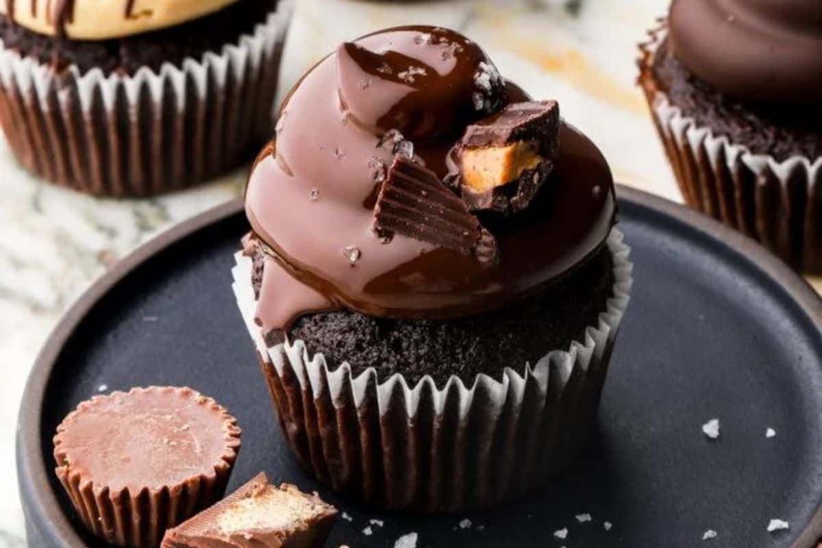 A chocolate cupcake topped with glossy chocolate frosting and pieces of peanut butter cups is displayed on a black plate. Bits of additional peanut butter cups are arranged next to the cupcake. Other similar cupcakes are blurred in the background.