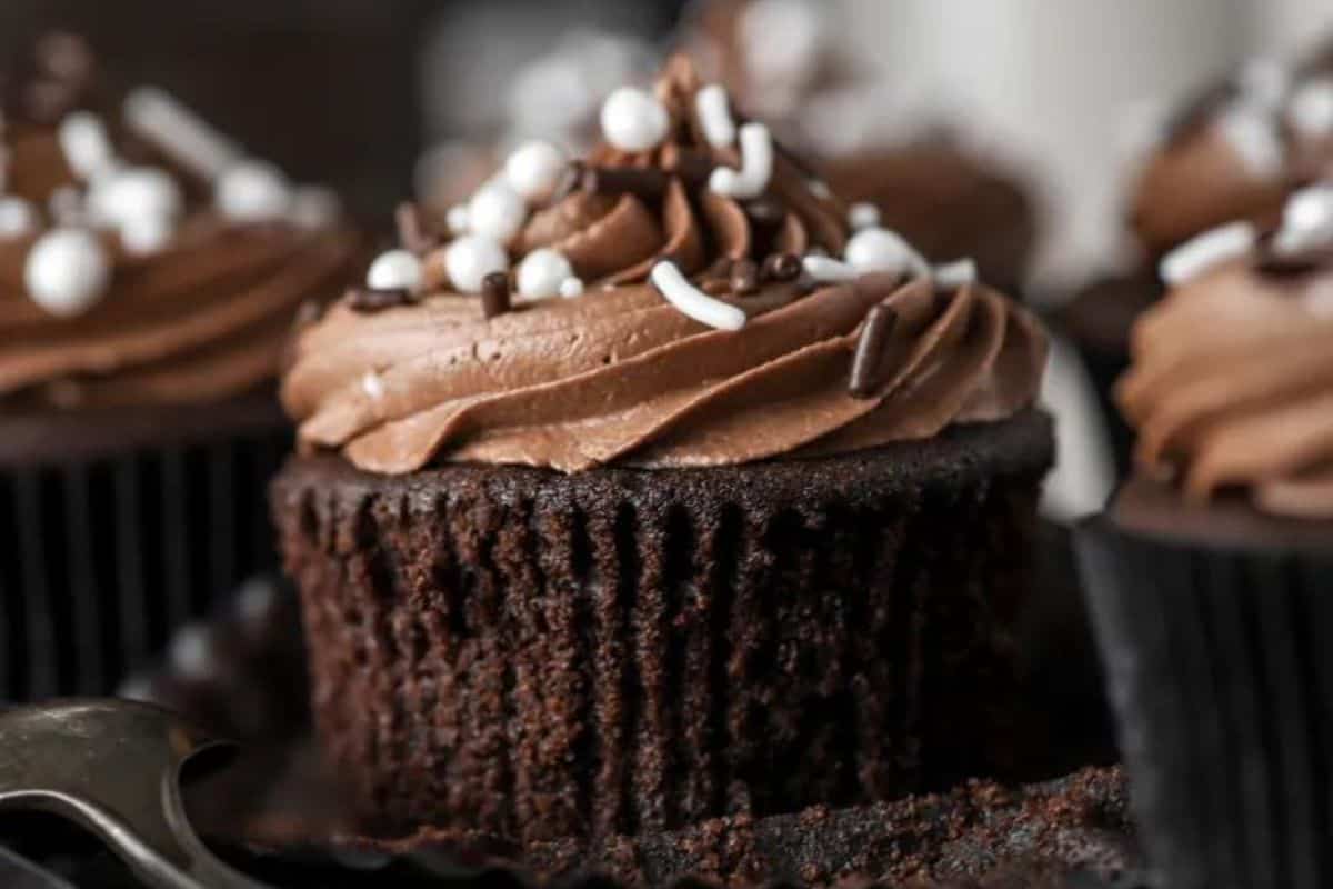 A close-up of a chocolate cupcake with swirled chocolate frosting, topped with white and chocolate sprinkles. The cupcake is in a dark wrapper, and other similarly decorated cupcakes are blurred in the background.