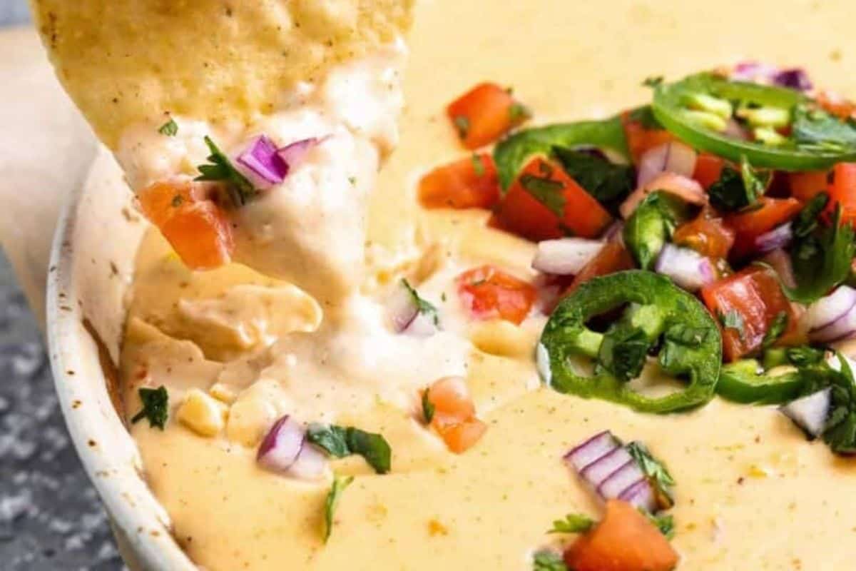 A close-up of a bowl of creamy queso dip being scooped by a tortilla chip. The queso is garnished with chopped red onions, tomatoes, cilantro, and slices of jalapeño, adding vibrant colors and textures to the creamy dip.