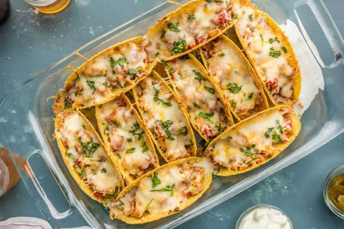 A glass baking dish holds several baked tacos, topped with melted cheese and garnished with fresh herbs. The tacos are neatly arranged, and surrounding the dish are bowls containing sour cream and other condiments.