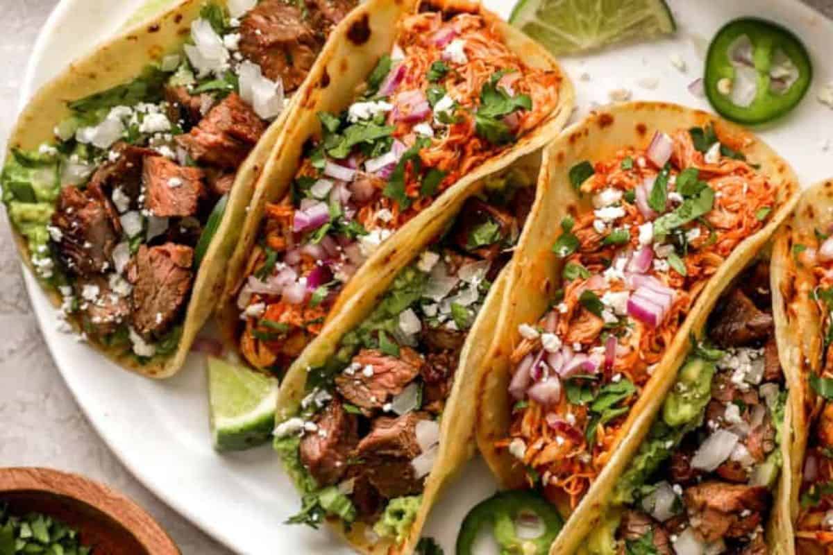 A plate of four tacos filled with chopped grilled meat, shredded cheese, diced red onions, chopped cilantro, jalapeño slices, and guacamole, all held in soft tortillas. Lime wedges are placed around the tacos as garnish.