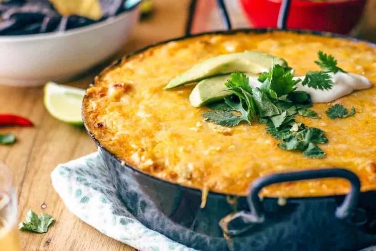 A baked dish topped with melted cheese, avocado slices, sour cream, and fresh cilantro. The black skillet rests on a patterned cloth, surrounded by lime wedges and a white bowl with tortilla chips in the background.