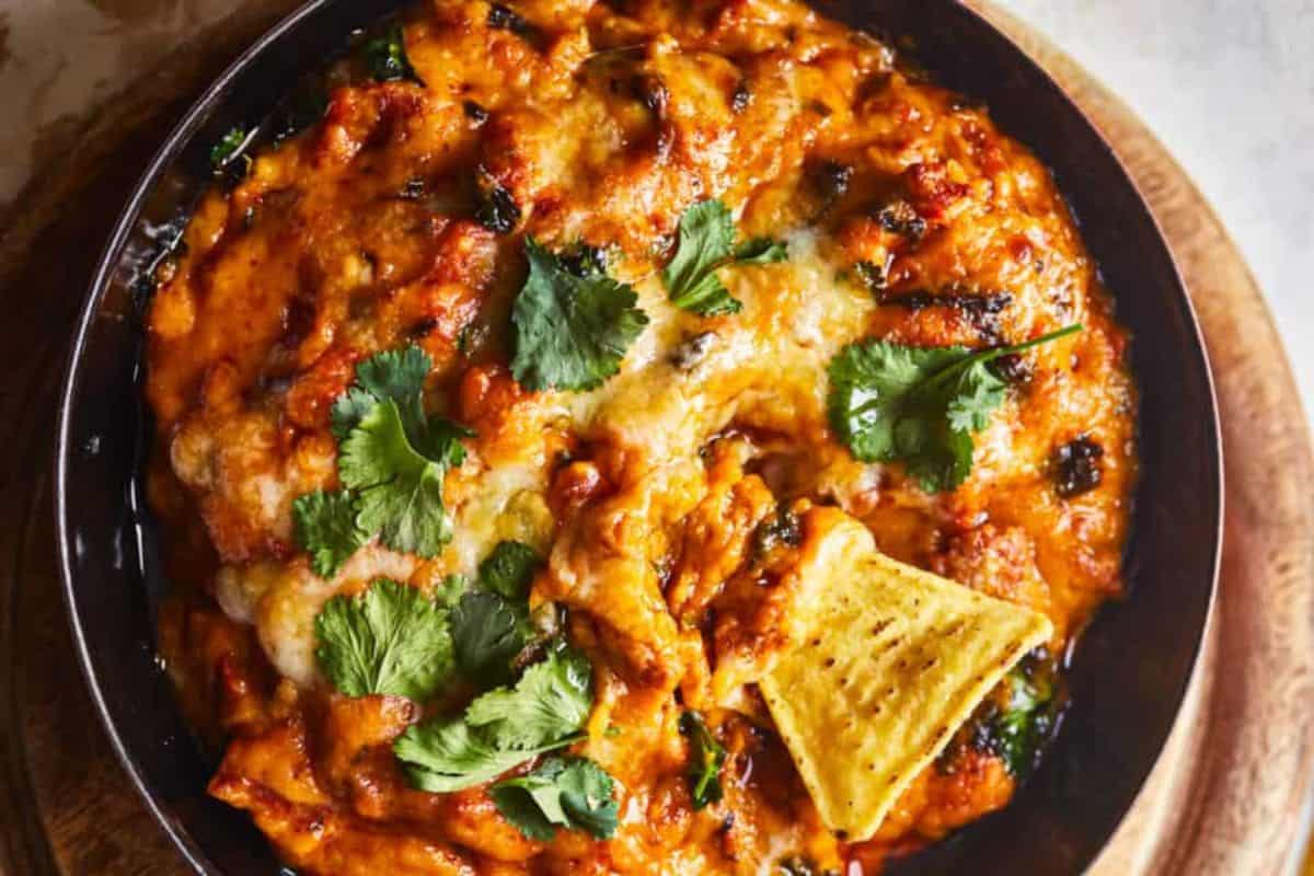 A skillet filled with baked dip topped with melted cheese, fresh cilantro leaves, and a single tortilla chip partially submerged near the center, placed on a wooden surface.