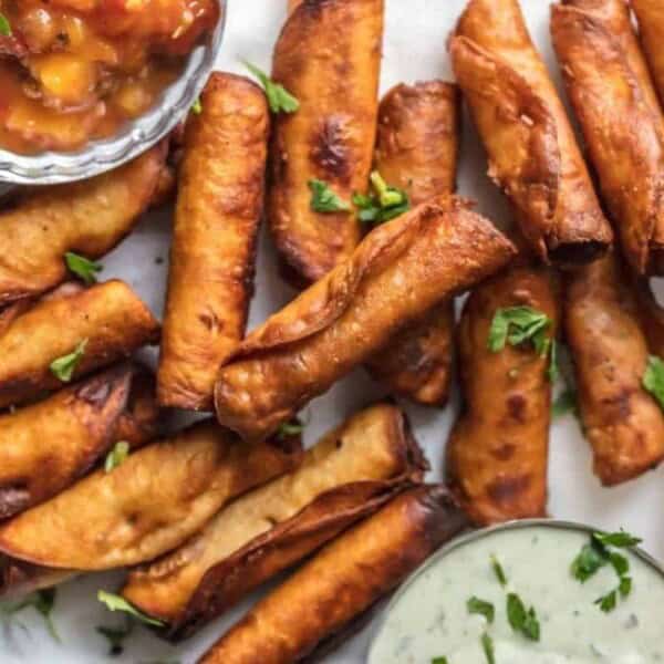 A plate filled with crispy, golden brown taquitos garnished with chopped fresh herbs. The taquitos are accompanied by small bowls of salsa and creamy dipping sauce, all placed on a white parchment paper-lined surface.