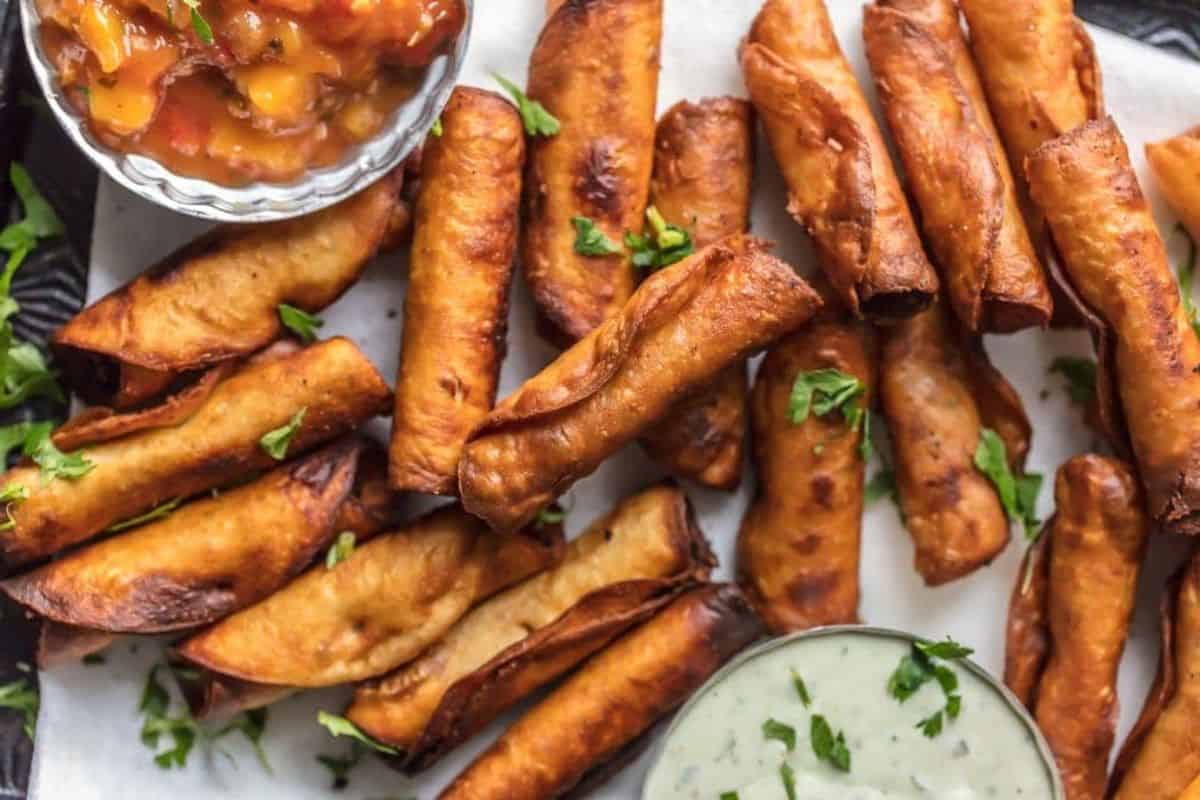 A plate filled with crispy, golden brown taquitos garnished with chopped fresh herbs. The taquitos are accompanied by small bowls of salsa and creamy dipping sauce, all placed on a white parchment paper-lined surface.