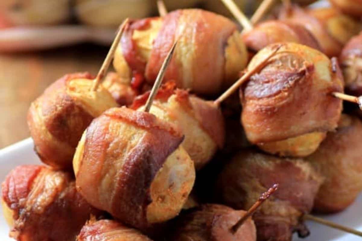 Close-up of bacon-wrapped water chestnuts on a white plate, secured with toothpicks. The water chestnuts are golden brown and crispy, with the bacon slightly caramelized, creating a savory appetizer.