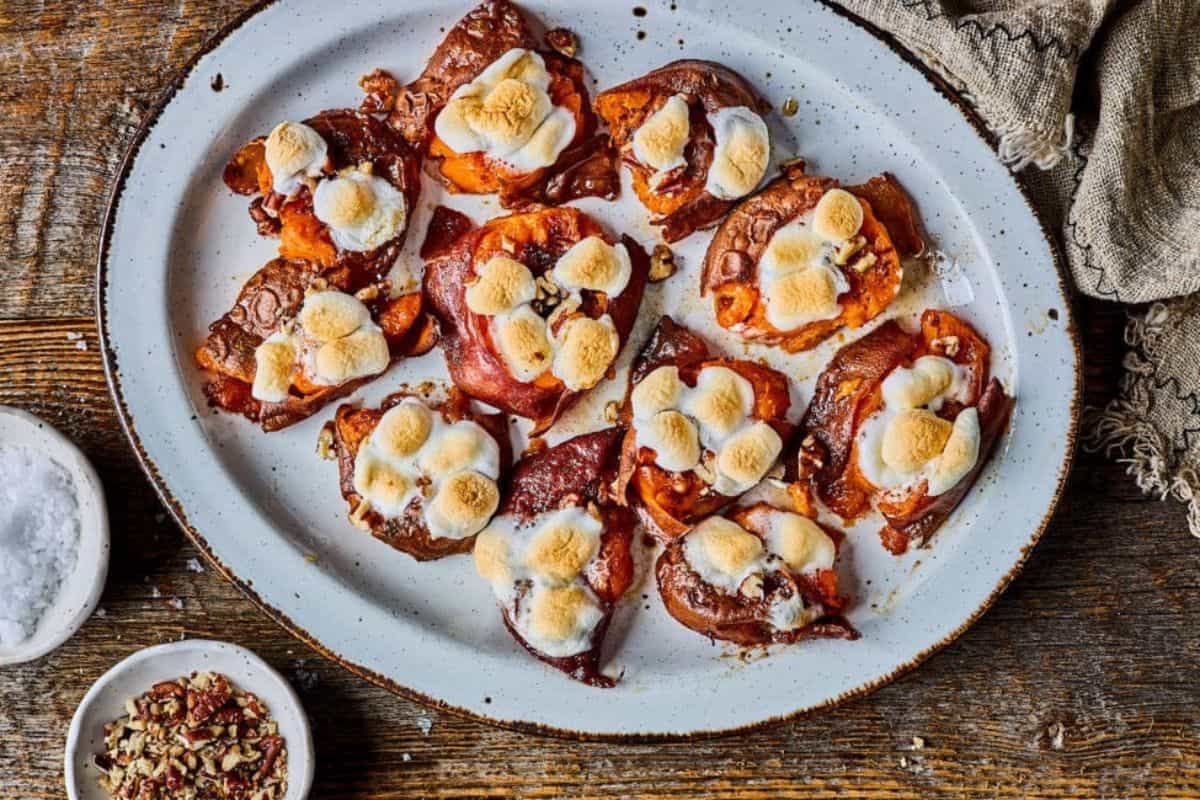 A white, oval plate with charred sweet potato rounds, each topped with toasted marshmallows and a sprinkle of chopped nuts. The plate is on a wooden table with a beige cloth napkin, a small dish of coarse salt, and a bowl of red pepper flakes.