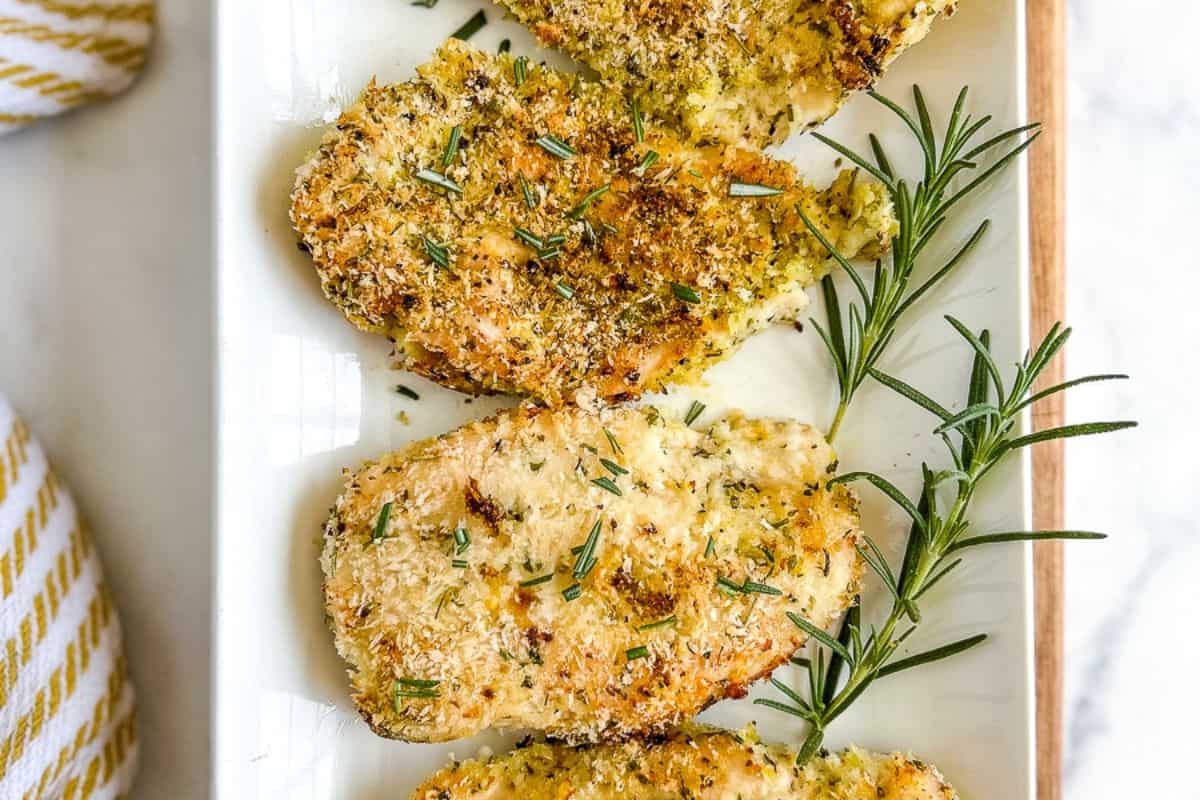 A rectangular white plate with four breaded and baked chicken breasts garnished with fresh rosemary sprigs. The chicken is golden brown and seasoned with herbs. The plate is partially placed on a white marble surface next to a yellow and white striped cloth.