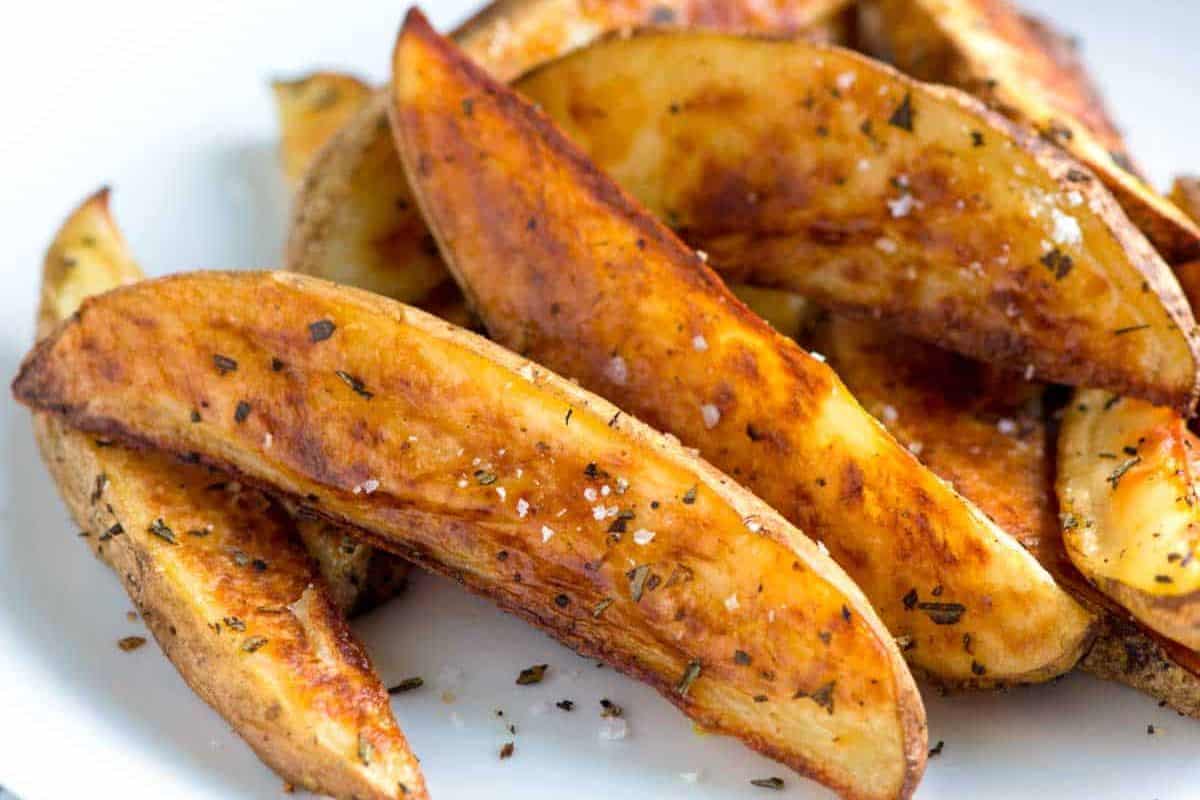 A close-up of seasoned potato wedges piled on a plate. The wedges are golden brown with crispy edges and sprinkled with herbs and coarse salt, showcasing a flavorful and appetizing appearance.