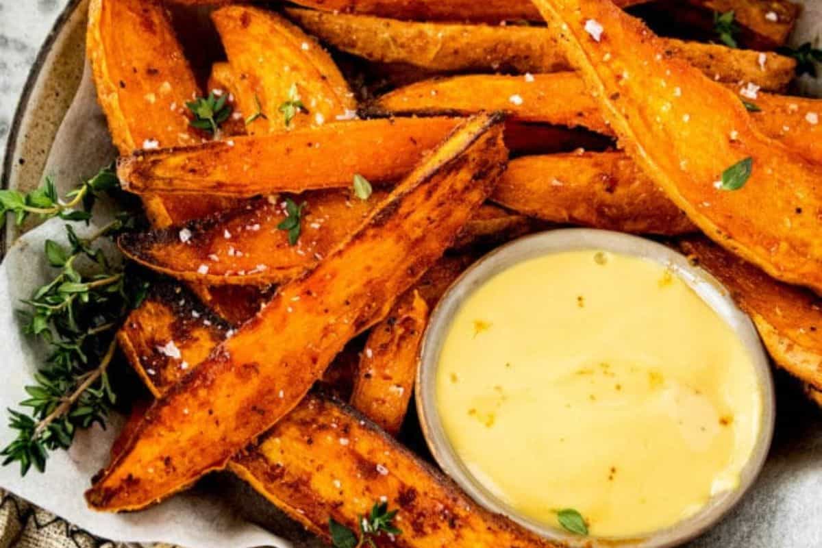 A plate of crispy, seasoned sweet potato wedges garnished with herbs is arranged next to a small bowl of creamy dipping sauce. The wedges are lightly sprinkled with coarse salt and fresh herbs.