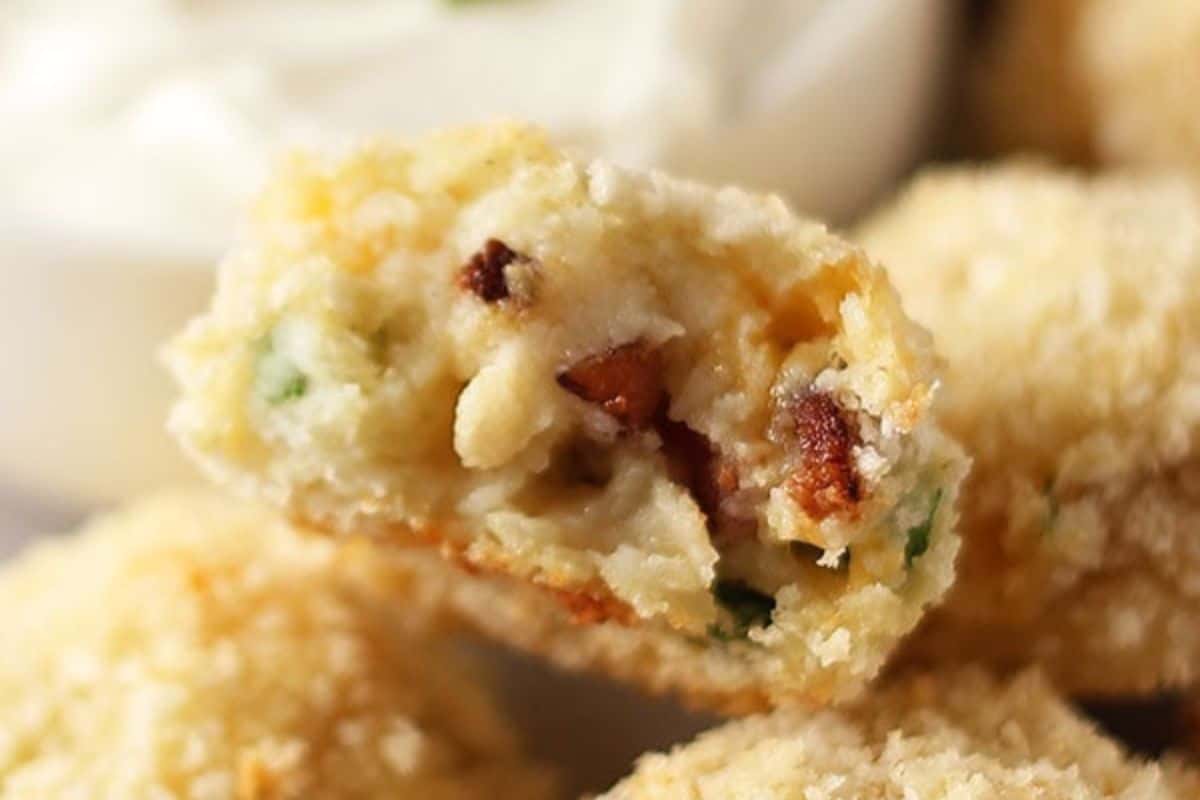 Close-up of a cut-open, crispy breaded and fried snack. The filling appears to contain small pieces of bacon or a similar meat, green herbs, and cheese. A dish of creamy dipping sauce is visible in the blurred background.