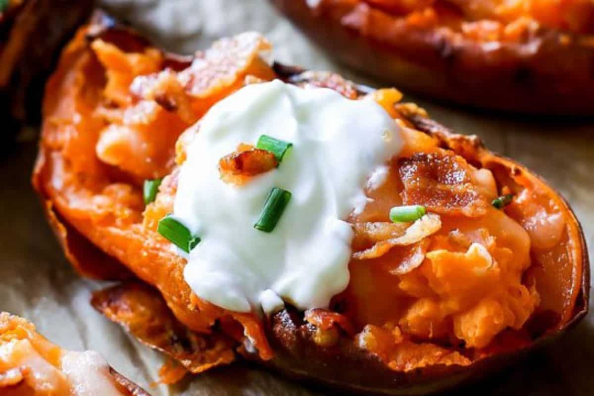 A close-up of a loaded baked sweet potato, topped with a dollop of sour cream, crispy bacon bits, and chopped green chives. The filling is a creamy mix of sweet potato and cheese. The skin is slightly crispy and browned.