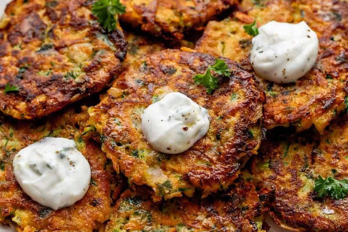 A close-up view of several golden-brown zucchini fritters, each topped with a dollop of sour cream and garnished with a small sprig of parsley. The fritters have a crispy texture, indicating they are freshly cooked.