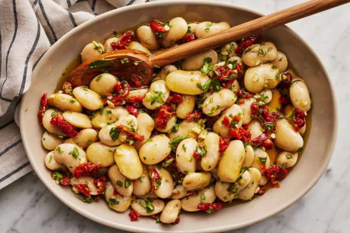 A bowl of marinated butter beans mixed with sun-dried tomatoes, chopped herbs, and olive oil. A wooden spoon rests inside the bowl. A striped cloth napkin is partially visible on the side. The dish is on a light-colored marble surface.