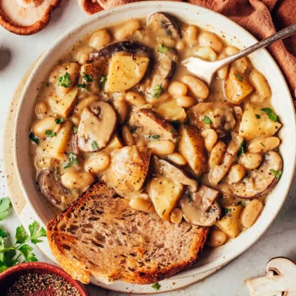 A hearty bowl of creamy mushroom and potato soup filled with white beans and garnished with fresh parsley. Two pieces of toasted bread are placed on the side of the bowl. Surrounding the bowl are fresh mushrooms, parsley, and a bowl of ground pepper.