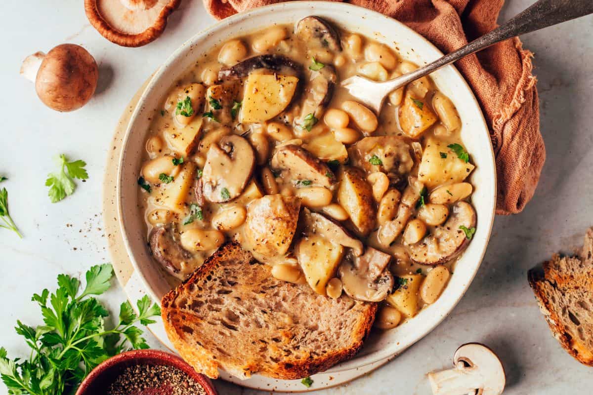 A hearty bowl of creamy mushroom and potato soup filled with white beans and garnished with fresh parsley. Two pieces of toasted bread are placed on the side of the bowl. Surrounding the bowl are fresh mushrooms, parsley, and a bowl of ground pepper.