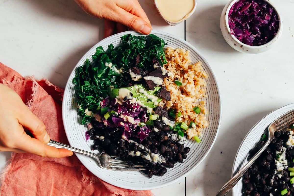 A vibrant meal bowl containing black beans, brown rice, greens, purple cabbage, and sliced avocado is being held by two hands. A creamy dressing is being poured on top. A small dish of shredded purple cabbage sits nearby on a white marble surface.