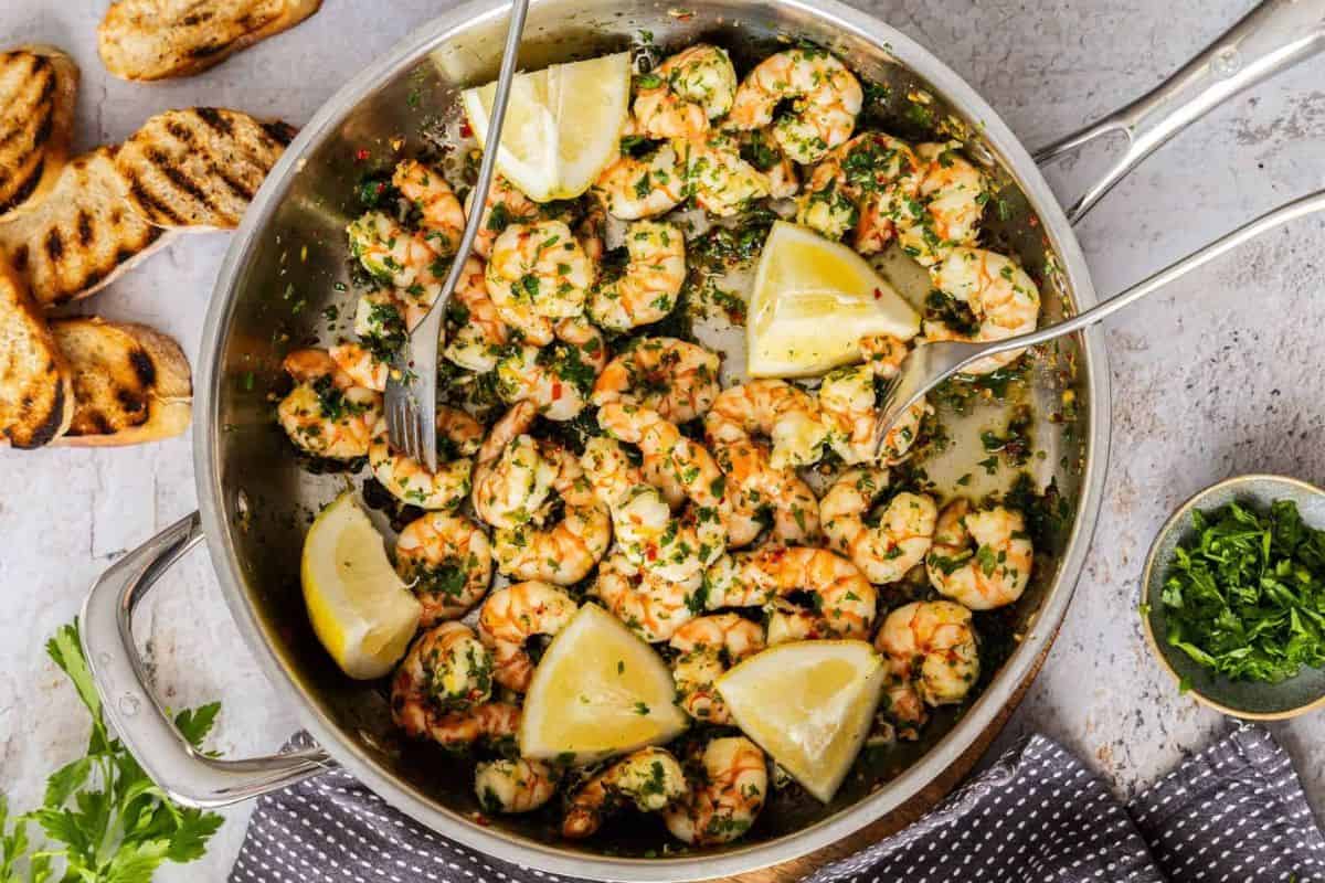 A skillet filled with cooked shrimp garnished with chopped herbs and lemon wedges. Two silver forks are positioned in the skillet. Grilled bread slices are on the side, and a small bowl of chopped herbs and a sprig of parsley are nearby.