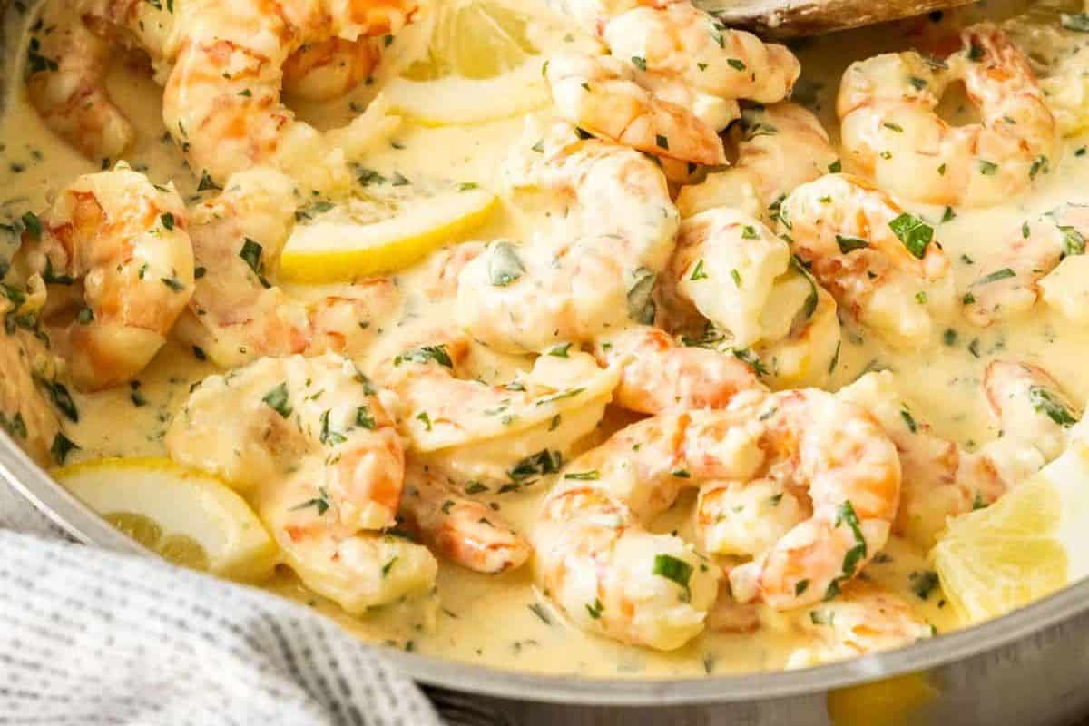 A close-up image of creamy garlic lemon shrimp in a pan. The dish features succulent shrimp bathed in a rich, creamy sauce, garnished with chopped parsley and fresh lemon slices for added zest. A wooden spoon is partially visible in the pan.