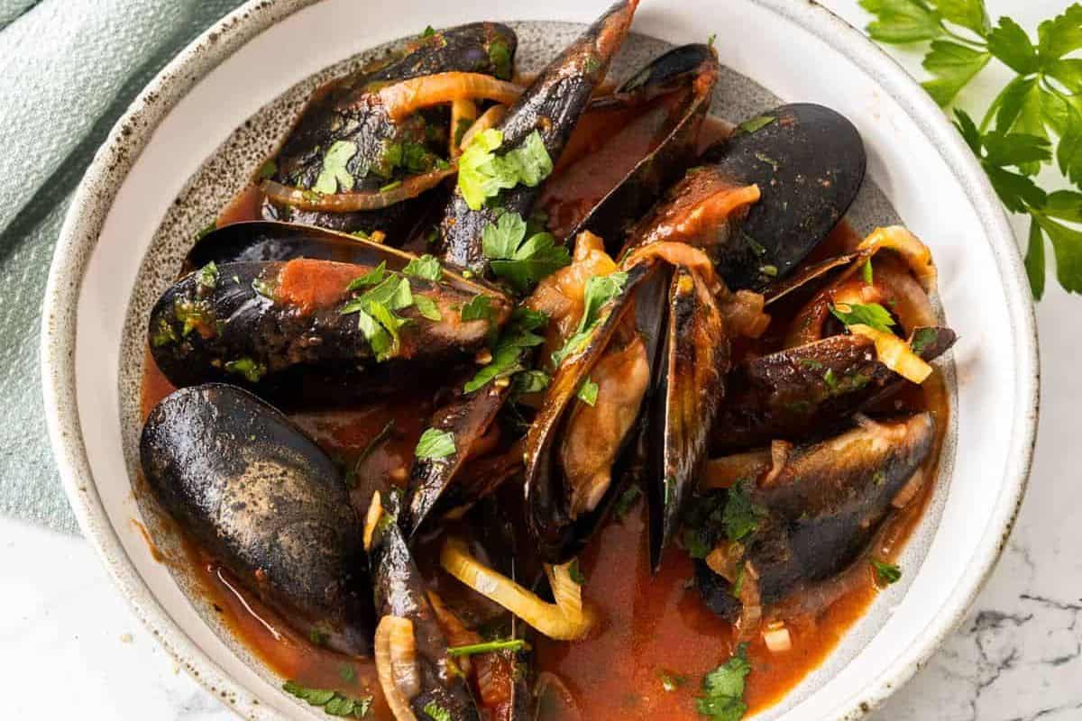 A bowl of mussels in a rich, red tomato sauce garnished with fresh parsley. The mussels are open, revealing their tender meat, and are surrounded by slices of onion and herbs. The dish is served in a white bowl on a marble surface with a sprig of parsley nearby.