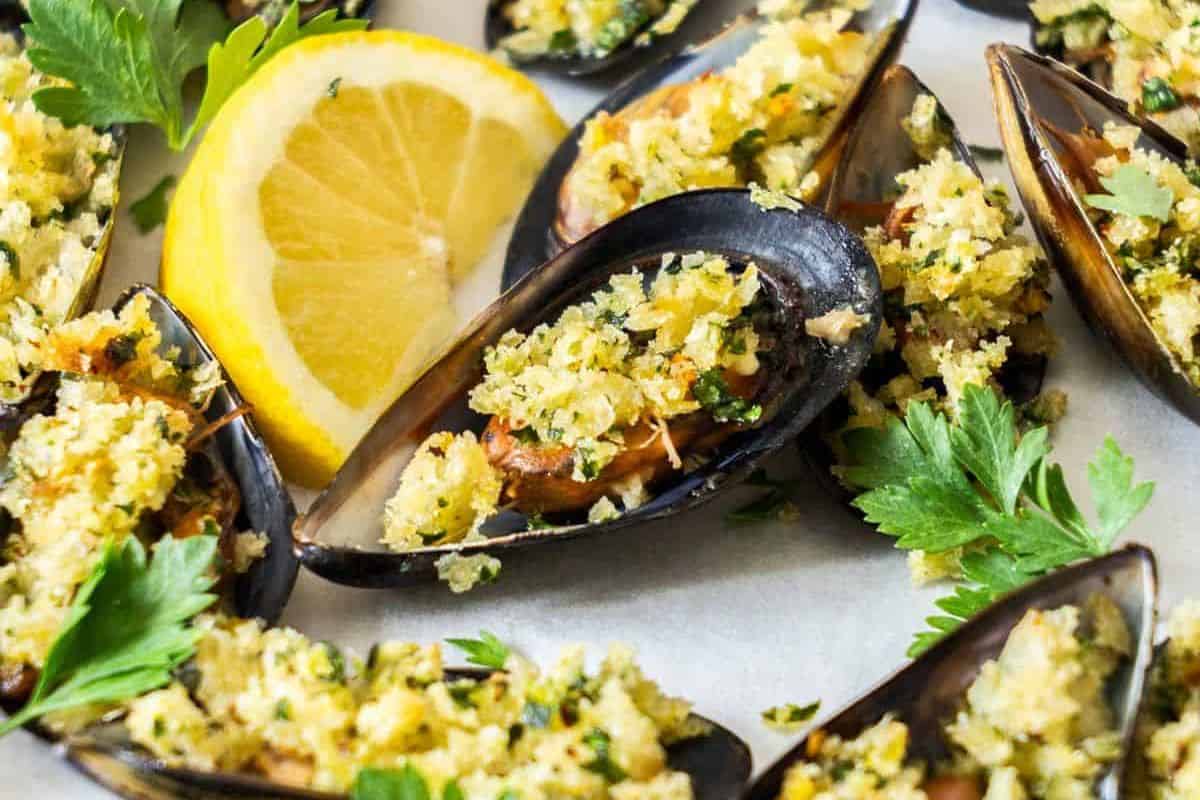 A close-up of baked mussels served on a plate. The mussels are topped with a golden breadcrumb and herb mixture, and garnished with fresh parsley. A slice of lemon is placed in the background for added freshness.