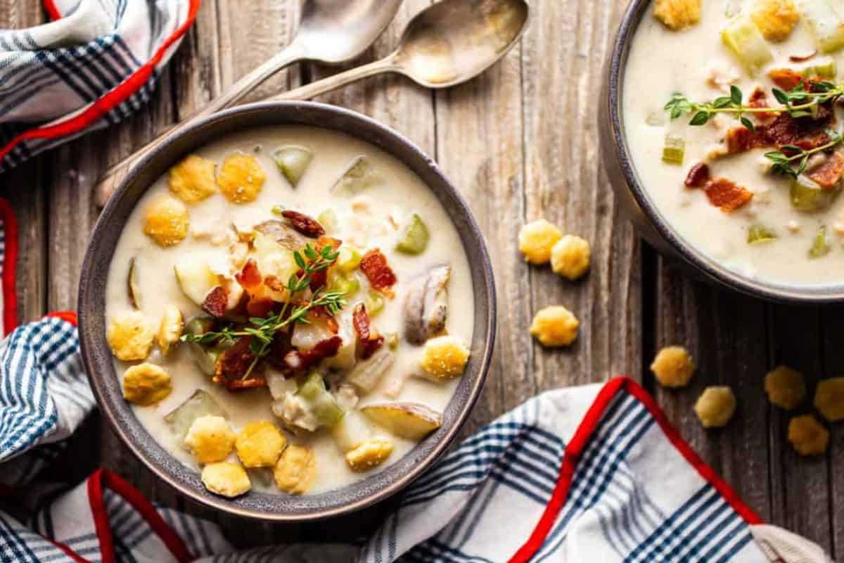 Two bowls of creamy clam chowder garnished with fresh herbs and crumbled bacon sit on a rustic wooden table. The soup is topped with oyster crackers, and a red, white, and blue checkered cloth and two spoons are in the background.