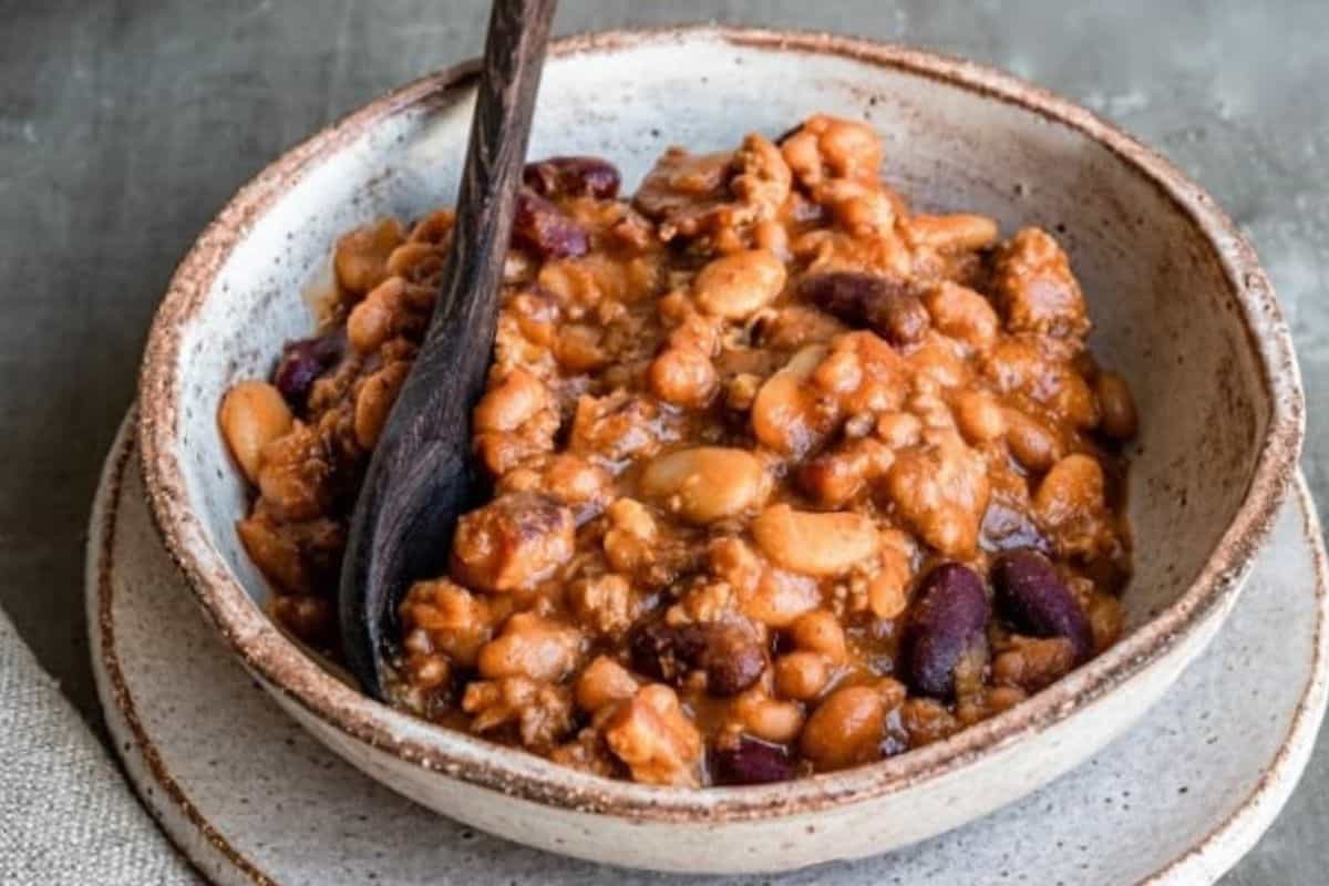 A rustic ceramic bowl filled with a hearty mixture of baked beans, featuring a variety of beans in a rich sauce. A dark wooden spoon is placed inside the bowl, and the bowl rests on a matching plate. The background is a neutral-toned surface.