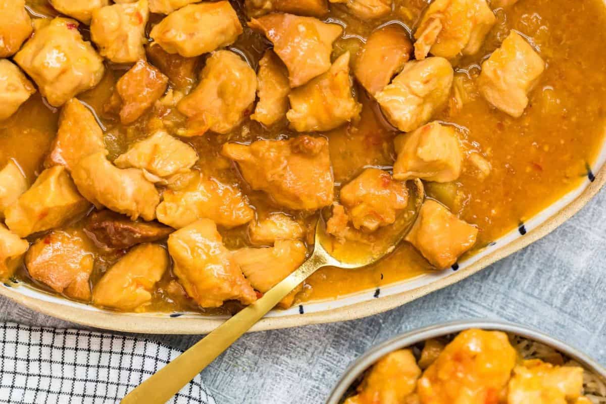 A dish featuring chunks of chicken in a rich, golden sauce is displayed in a large serving dish with a spoon. The dish is placed on a checkered cloth, with a small bowl containing a serving of the same chicken dish in the foreground.