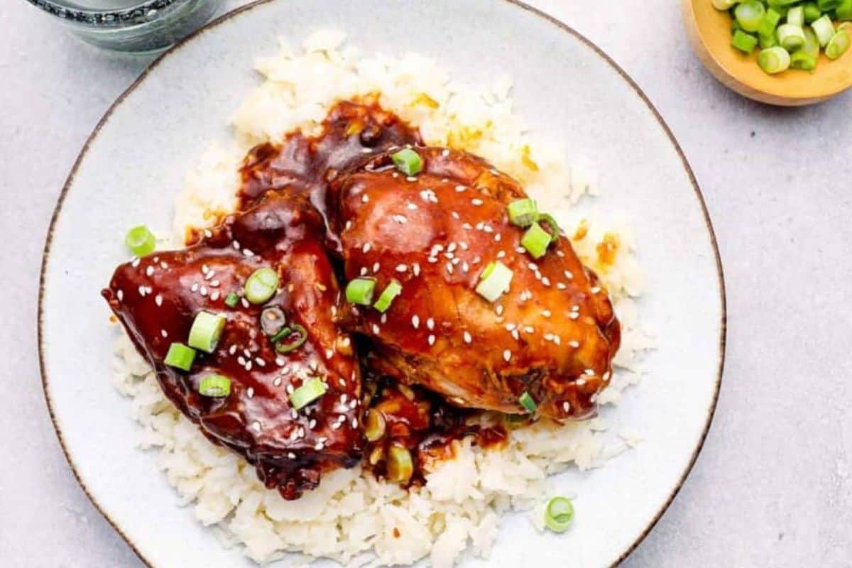 A plate with two pieces of glistening teriyaki chicken topped with white sesame seeds and chopped green onions, served on a bed of white rice. A small bowl of additional chopped green onions is visible in the background.