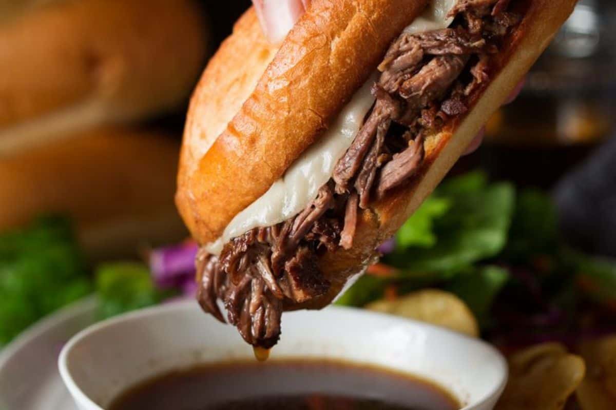 Close-up of a hand dipping a French dip sandwich with shredded beef and melted cheese into a bowl of au jus. The background shows part of the sandwich and a blurred salad with leafy greens.