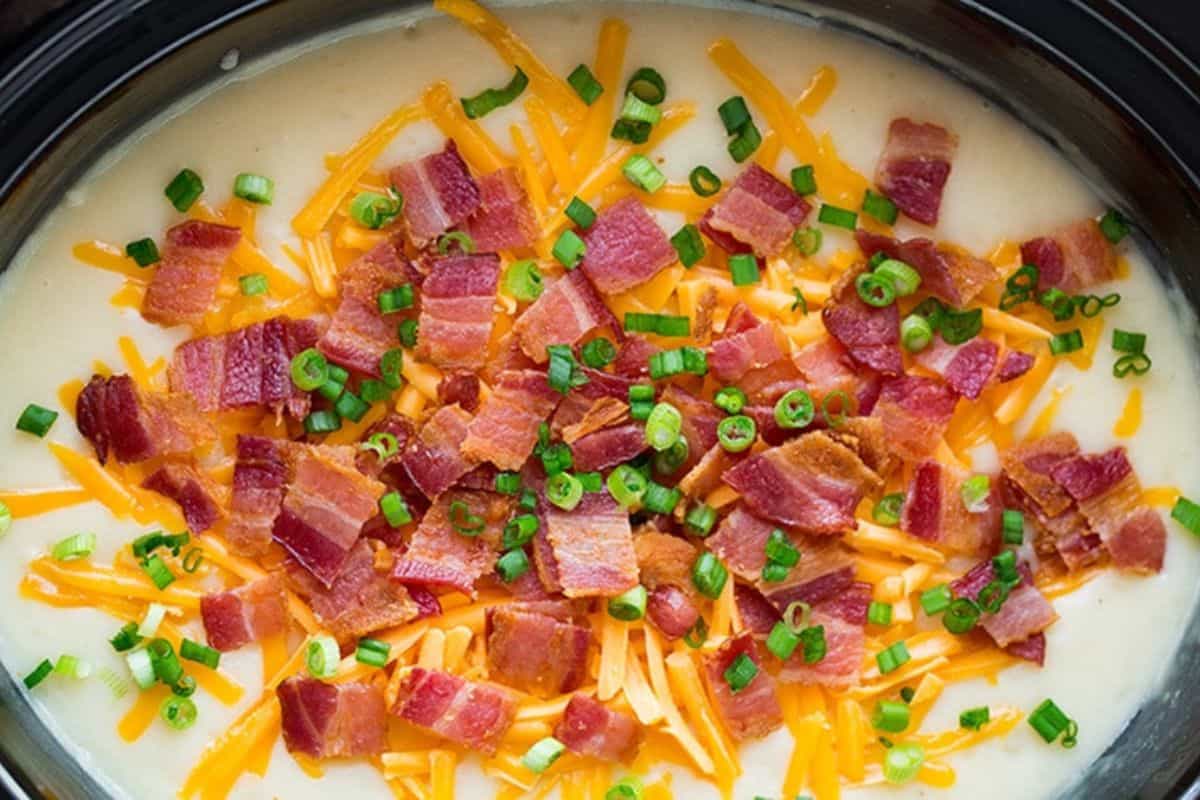 A creamy soup in a slow cooker, topped generously with shredded cheddar cheese, crispy bacon pieces, and chopped green onions.