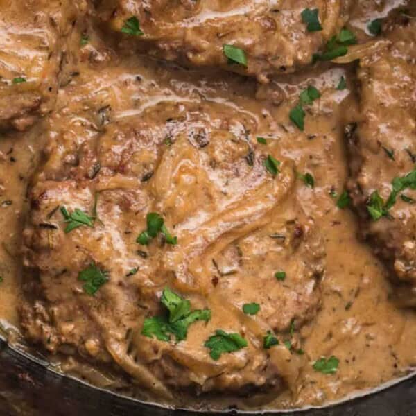Close-up of Salisbury steaks covered in creamy mushroom and onion gravy, garnished with chopped parsley. The dish appears savory with a rich, thick sauce coating the beef patties and bits of cooked onions visible throughout.